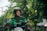A person wearing a helmet and a green jacket with a Grab logo is riding a motorcycle. The background features a blurred mix of greenery and urban elements, suggesting an outdoor setting.
