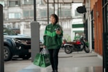 A person wearing a green Grab jacket walks outside an urban setting, holding a smartphone and carrying Grab Food delivery bags. The background includes parked vehicles and a building with large windows.