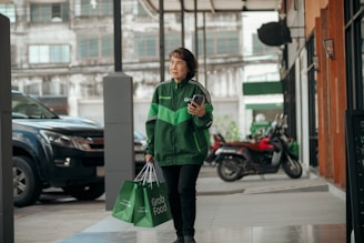 A person wearing a green Grab jacket walks outside an urban setting, holding a smartphone and carrying Grab Food delivery bags. The background includes parked vehicles and a building with large windows.