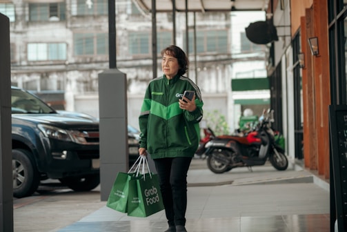 A person wearing a green Grab jacket walks outside an urban setting, holding a smartphone and carrying Grab Food delivery bags. The background includes parked vehicles and a building with large windows.