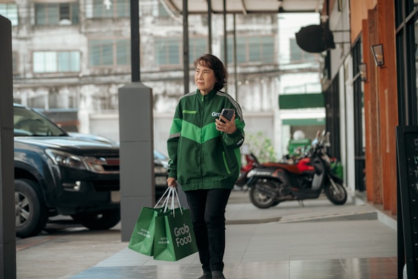 A person wearing a green Grab jacket walks outside an urban setting, holding a smartphone and carrying Grab Food delivery bags. The background includes parked vehicles and a building with large windows.