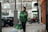 A person wearing a green Grab jacket walks outside an urban setting, holding a smartphone and carrying Grab Food delivery bags. The background includes parked vehicles and a building with large windows.