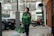 A person wearing a green Grab jacket walks outside an urban setting, holding a smartphone and carrying Grab Food delivery bags. The background includes parked vehicles and a building with large windows.