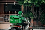 A delivery person wearing a green uniform and helmet rides a scooter with a large green delivery box on the back. The logo 'Grab' is visible on the box and the uniform. The setting appears to be a residential area with a gate, plants, and trees visible in the background.