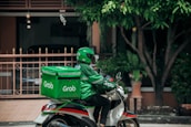 A delivery person wearing a green uniform and helmet rides a scooter with a large green delivery box on the back. The logo 'Grab' is visible on the box and the uniform. The setting appears to be a residential area with a gate, plants, and trees visible in the background.