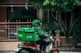 A delivery person wearing a green uniform and helmet rides a scooter with a large green delivery box on the back. The logo 'Grab' is visible on the box and the uniform. The setting appears to be a residential area with a gate, plants, and trees visible in the background.