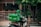 A delivery person wearing a green uniform and helmet rides a scooter with a large green delivery box on the back. The logo 'Grab' is visible on the box and the uniform. The setting appears to be a residential area with a gate, plants, and trees visible in the background.