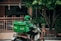 A delivery person wearing a green uniform and helmet rides a scooter with a large green delivery box on the back. The logo 'Grab' is visible on the box and the uniform. The setting appears to be a residential area with a gate, plants, and trees visible in the background.