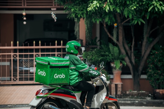 A delivery person wearing a green uniform and helmet rides a scooter with a large green delivery box on the back. The logo 'Grab' is visible on the box and the uniform. The setting appears to be a residential area with a gate, plants, and trees visible in the background.
