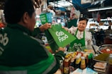 A vendor in a bustling market hands a GrabFood delivery bag to a person wearing a GrabFood uniform. The stall is filled with various jars and bottles, suggesting it sells condiments or sauces. The vendor is smiling, and the setting is lively with numerous products displayed around the stall.