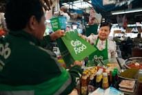 A vendor in a bustling market hands a GrabFood delivery bag to a person wearing a GrabFood uniform. The stall is filled with various jars and bottles, suggesting it sells condiments or sauces. The vendor is smiling, and the setting is lively with numerous products displayed around the stall.