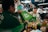 A vendor in a bustling market hands a GrabFood delivery bag to a person wearing a GrabFood uniform. The stall is filled with various jars and bottles, suggesting it sells condiments or sauces. The vendor is smiling, and the setting is lively with numerous products displayed around the stall.