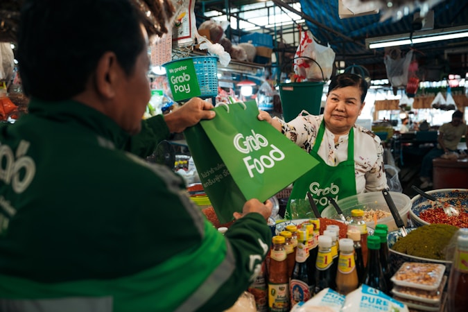 A vendor in a bustling market hands a GrabFood delivery bag to a person wearing a GrabFood uniform. The stall is filled with various jars and bottles, suggesting it sells condiments or sauces. The vendor is smiling, and the setting is lively with numerous products displayed around the stall.