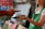 A person wearing a green apron holds a smartphone in their hand while another person extends a hand with a different smartphone, possibly indicating an exchange or payment process using mobile devices. The background appears to be a blurred market or shop setting.