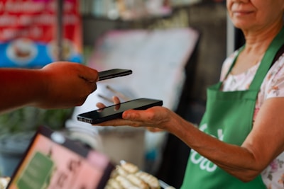 A person wearing a green apron holds a smartphone in their hand while another person extends a hand with a different smartphone, possibly indicating an exchange or payment process using mobile devices. The background appears to be a blurred market or shop setting.