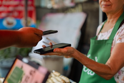 A person wearing a green apron holds a smartphone in their hand while another person extends a hand with a different smartphone, possibly indicating an exchange or payment process using mobile devices. The background appears to be a blurred market or shop setting.