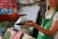 A person wearing a green apron holds a smartphone in their hand while another person extends a hand with a different smartphone, possibly indicating an exchange or payment process using mobile devices. The background appears to be a blurred market or shop setting.