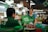 A woman in a market stall is handing a green Grab Food bag to a man wearing a green jacket with the Grab logo. The market stall has various fresh produce including baskets of chilies and other vegetables. There are signs indicating the acceptance of Grab Pay and other digital payments.