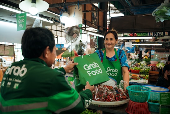 A woman in a market stall is handing a green Grab Food bag to a man wearing a green jacket with the Grab logo. The market stall has various fresh produce including baskets of chilies and other vegetables. There are signs indicating the acceptance of Grab Pay and other digital payments.