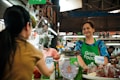 A market vendor wearing a green 'Grab Food' apron is smiling behind a stall with various vegetables and spices. A customer is scanning a QR code with a smartphone. The setting has a vibrant and lively atmosphere typical of local markets.