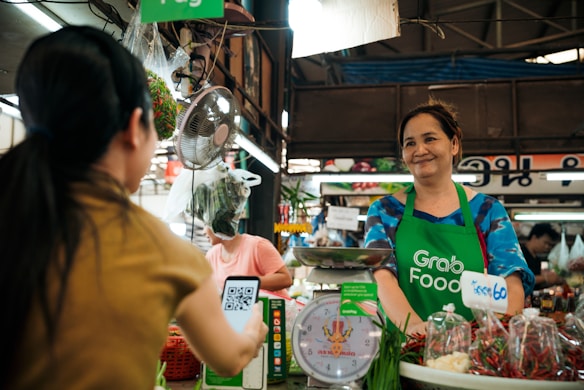 A market vendor wearing a green 'Grab Food' apron is smiling behind a stall with various vegetables and spices. A customer is scanning a QR code with a smartphone. The setting has a vibrant and lively atmosphere typical of local markets.