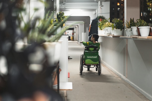 A person using a wheelchair is seen moving down a long corridor lined with potted plants. The individual is wearing a green uniform, possibly from a delivery service, with a large GrabFood bag on the back of the wheelchair. The corridor is brightly lit, with a focus on the greenery and the person in motion.