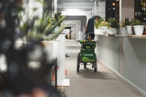 A person using a wheelchair is seen moving down a long corridor lined with potted plants. The individual is wearing a green uniform, possibly from a delivery service, with a large GrabFood bag on the back of the wheelchair. The corridor is brightly lit, with a focus on the greenery and the person in motion.