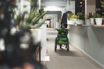 A person using a wheelchair is seen moving down a long corridor lined with potted plants. The individual is wearing a green uniform, possibly from a delivery service, with a large GrabFood bag on the back of the wheelchair. The corridor is brightly lit, with a focus on the greenery and the person in motion.