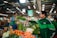 A vibrant market setting features a vendor and a customer in exchange. The vendor, smiling warmly, hands over a basket filled with fresh greens. The market stall is abundant with various vegetables such as tomatoes, carrots, and radishes, neatly displayed. Overhead lighting casts a bright, inviting glow, and signage suggests digital payment options.