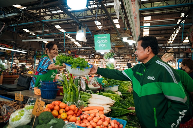 A vibrant market setting features a vendor and a customer in exchange. The vendor, smiling warmly, hands over a basket filled with fresh greens. The market stall is abundant with various vegetables such as tomatoes, carrots, and radishes, neatly displayed. Overhead lighting casts a bright, inviting glow, and signage suggests digital payment options.