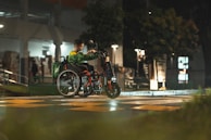 A person in a green jacket sits in a wheelchair that is customized with an electric attachment at the front, crossing a street at night. The scene is dimly lit with streetlights reflecting off the wet pavement, and there are blurred trees and buildings in the background.