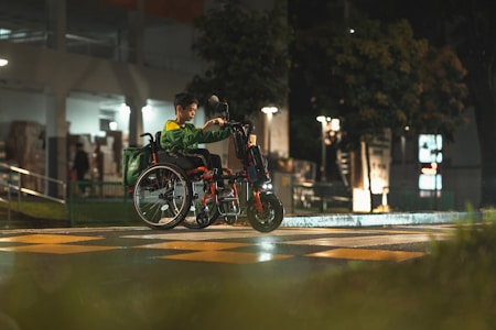 A person in a green jacket sits in a wheelchair that is customized with an electric attachment at the front, crossing a street at night. The scene is dimly lit with streetlights reflecting off the wet pavement, and there are blurred trees and buildings in the background.
