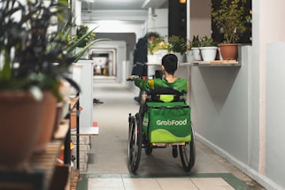 A person wearing a green uniform with a 'GrabFood' backpack is seated in a wheelchair moving down a corridor. The corridor is lined with plants in pots on both sides, and there is another individual walking further ahead.