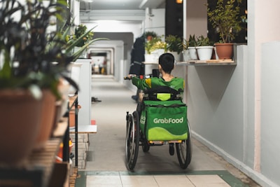 A person wearing a green uniform with a 'GrabFood' backpack is seated in a wheelchair moving down a corridor. The corridor is lined with plants in pots on both sides, and there is another individual walking further ahead.