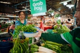 A woman in a green apron is handing a basket of greens to a customer at a bustling market. Various fresh vegetables, including onions, chilies, and leafy greens, are displayed on the stall. The environment is vibrant with shoppers and vendors engaged in transactions. Bright overhead lights illuminate the scene, and a sign promoting cashless payment is visible.