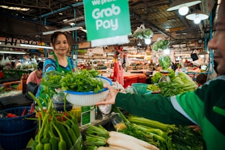 A woman in a green apron is handing a basket of greens to a customer at a bustling market. Various fresh vegetables, including onions, chilies, and leafy greens, are displayed on the stall. The environment is vibrant with shoppers and vendors engaged in transactions. Bright overhead lights illuminate the scene, and a sign promoting cashless payment is visible.