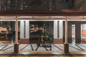 A person in a green jacket sits in a wheelchair under a covered walkway. It is nighttime, and the surroundings include residential buildings and parked vehicles. The area is wet, suggesting recent rain. The ground features a geometric pattern in shades of gray and brown.
