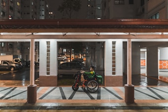 A person in a green jacket sits in a wheelchair under a covered walkway. It is nighttime, and the surroundings include residential buildings and parked vehicles. The area is wet, suggesting recent rain. The ground features a geometric pattern in shades of gray and brown.