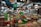 A vendor wears a bright green Grab Food apron while serving products in a bustling market. There are a variety of items on display, including jars, bottles of sauces, spices in bowls, and hanging bags. The market is filled with diverse goods and has a lively atmosphere.
