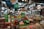 A vendor wears a bright green Grab Food apron while serving products in a bustling market. There are a variety of items on display, including jars, bottles of sauces, spices in bowls, and hanging bags. The market is filled with diverse goods and has a lively atmosphere.
