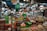 A vendor wears a bright green Grab Food apron while serving products in a bustling market. There are a variety of items on display, including jars, bottles of sauces, spices in bowls, and hanging bags. The market is filled with diverse goods and has a lively atmosphere.