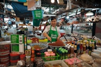 A vendor wears a bright green Grab Food apron while serving products in a bustling market. There are a variety of items on display, including jars, bottles of sauces, spices in bowls, and hanging bags. The market is filled with diverse goods and has a lively atmosphere.