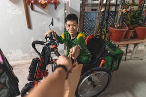 A young person in a green and yellow shirt smiles while reaching out to take a brown bag. They are on a tricycle adapted for use as a wheelchair. Several potted plants and a decorative wall hang in the background.