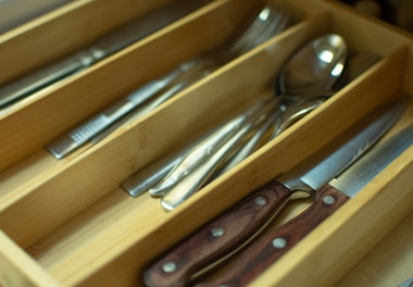 A wooden cutlery drawer organizer contains various silver utensils, including knives, forks, and spoons, neatly arranged in separate compartments. A knife with a wooden handle is prominently visible.