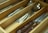A wooden cutlery drawer organizer contains various silver utensils, including knives, forks, and spoons, neatly arranged in separate compartments. A knife with a wooden handle is prominently visible.