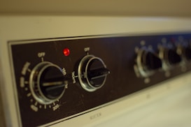 Close-up view of a kitchen appliance control panel featuring several black knobs with temperature settings visible and a small red indicator light. The panel has labels for various cooking modes such as 'Broil' and temperature increments, with the design emphasizing functionality.
