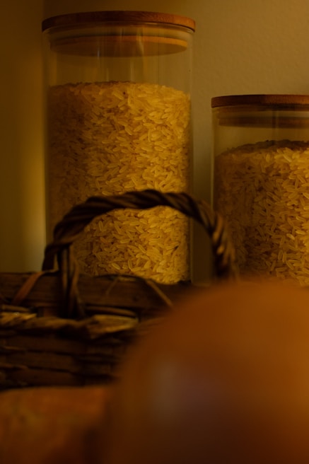 Two glass jars filled with uncooked rice are placed side by side, with wooden lids on top. A woven wicker basket with a handle is partially visible in the foreground.