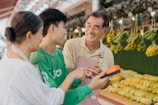Three people are interacting cheerfully at a fruit market. A vendor wearing an apron is smiling at two customers. Various fruits, including bananas and other fresh produce, are displayed in the background. One customer is holding a device, possibly a payment terminal.