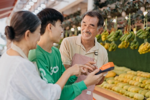 Three people are interacting cheerfully at a fruit market. A vendor wearing an apron is smiling at two customers. Various fruits, including bananas and other fresh produce, are displayed in the background. One customer is holding a device, possibly a payment terminal.