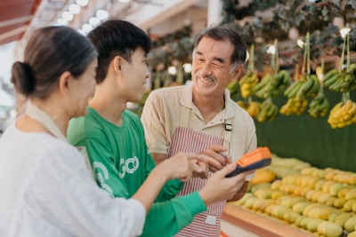 Three people are interacting cheerfully at a fruit market. A vendor wearing an apron is smiling at two customers. Various fruits, including bananas and other fresh produce, are displayed in the background. One customer is holding a device, possibly a payment terminal.